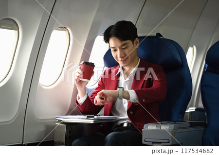 Young businessman holding cup of coffee and looking at his watch while sitting in an airplane seat 117534682
