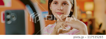 Gen alpha teen girl with arm prosthesis showing hand heart while smartphone camera on tripod recording video, website banner Gen alpha teen girl with arm prosthesis showing hand heart while smartphone camera on tripod recording video, website banner 117535265