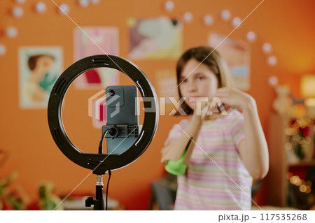Selective focus of teen girl with arm prosthesis posing while smartphone camera on tripod with LED lamp recording video 117535268