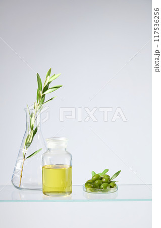 White minimal background photo in vertical frame for cosmetic advertising, which has olive ingredient. A glass bottle filled by olive oil, next to olives dish and erlenmeyer with a green olive branch 117536256