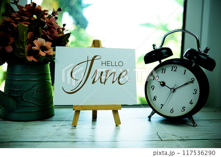 Hello June text on paper card with easel and alarm clock in the window background Hello June text on paper card with easel and alarm clock in the window background 117536290