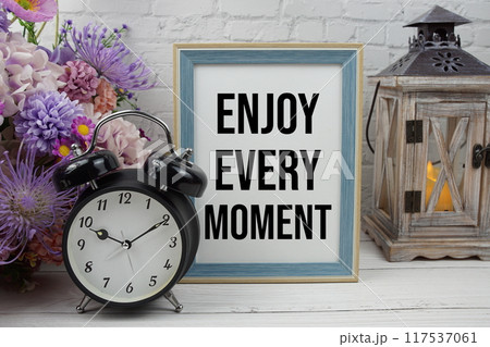 Enjoy every moment text message motivational and inspiration quote with flowers decoration on white brick wall and wooden background Enjoy every moment text message motivational and inspiration quote with flowers decoration on white brick wall and wooden background 117537061
