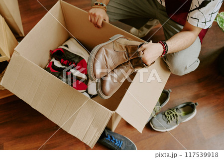 Person sorting various pairs of shoes in cardboard box on wooden floor. Different styles of shoes present, with some already inside box and others around it on floor Person sorting various pairs of shoes in cardboard box on wooden floor. Different styles of shoes present, with some already inside box and others around it on floor 117539918
