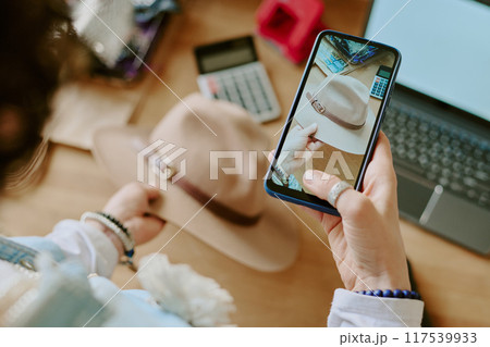 Person holding smartphone, capturing image of beige hat, with calculator and laptop on desk, focusing on detailed buckle. Scene set in warm, closely arranged workspace 117539933