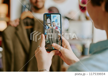 Person holding smartphone capturing video of another individual holding brown jacket in a store setting focusing on the clothing item 117539934