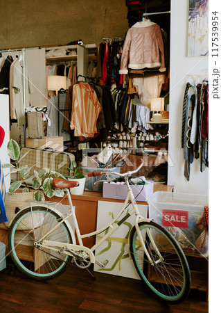 Scene depicting vintage bicycle and variety of clothing in store having sale sign. Clothing racks and accessories are visible in background enhancing cozy shop atmosphere 117539954