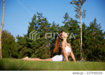 Young fit woman, in white fit wear doing yoga exercises, stretching on green grass at park against blurred background of forest. Young fit woman, in white fit wear doing yoga exercises, stretching on green grass at park against blurred background of forest. 117540326
