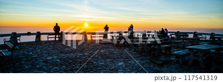 A group of individuals stands at a viewpoint, taking in the breathtaking view of the sunset. Warm hues paint the sky as the sun descends, creating a perfect moment for reflection and connection. 117541319