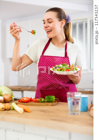 Vegeterian woman standing at table in kitchen and eating vegetable salad Vegeterian woman standing at table in kitchen and eating vegetable salad 117543237