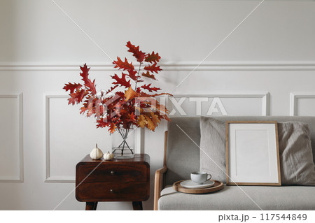 Red oak tree leaves, branches in vase on wooden night table. Linen midcentury sofa with cup of coffee. Blank wooden picture frame mockup. Little white pumpkins. Autumn interior. Thanksgiving 117544849
