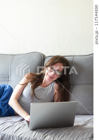 A young woman with glasses lies on the sofa and communicates via video conference via a laptop. A young woman with glasses lies on the sofa and communicates via video conference via a laptop. 117544890