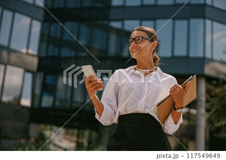 Confident businesswoman with smartphone and folder outside a modern office building 117549649