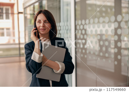 Professional Businesswoman Holding Laptop While Talking on Phone in Modern Office Setting 117549660