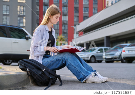 Young college student, girl with backpack reading textbook, outdoors Young college student, girl with backpack reading textbook, outdoors 117551687
