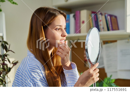 Young beautiful woman looking at face hair skin makeup teeth in mirror 117551691