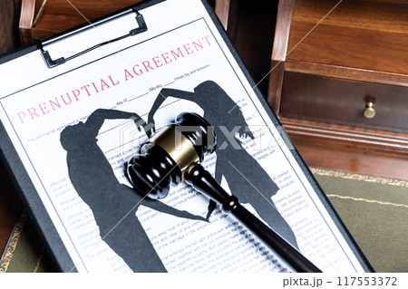 A clipboard holding a prenuptial agreement with a cut-out silhouette of a couple and wedding rings, next to a judge's gavel. 117553372