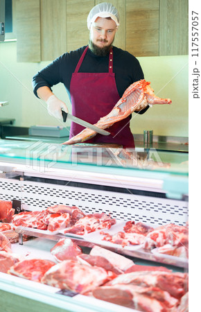 Skilled bearded butcher cutting fresh lamb carcass at counter 117557057