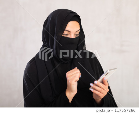 young positive girl with her face covered by burka holds phone in her hands and surf Internet 117557266
