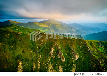 Breathtaking aerial view of green, rugged mountain peaks under dramatic sky. Scene captures contrast between vibrant landscape and moody clouds, with sunlight breaking through. Carpathians, Chornogora 117560677