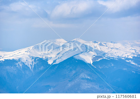 Snow-covered mountain peak under clear blue sky, with patches of snow on slopes and scattered trees. Serene winter landscape contrasts sharply with the dark, forested areas below. Blyznytsya, Ukraine 117560681