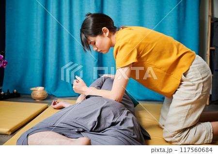 Masseuse making thai yoga massage. Woman therapist doing traditional massage treatment using elbow for male patient indoors. Client lying on mat, meditating, enjoying hand massage in calm atmosphere. Masseuse making thai yoga massage. Woman therapist doing traditional massage treatment using elbow for male patient indoors. Client lying on mat, meditating, enjoying hand massage in calm atmosphere. 117560684