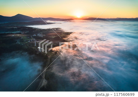 Stunning aerial view of sunrise over mountainous landscape, with thick fog blanketing valleys and hills. Sun's golden rays pierce through mist, casting serene glow over the entire scene. 117560699