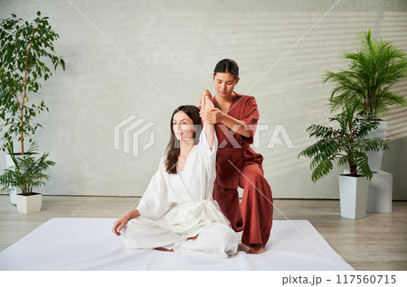 Masseuse making thai yoga massage. Woman therapist doing traditional massage treatment for female patient indoors. Client sitting on mat, meditating, enjoying hand massage in calm atmosphere. 117560715