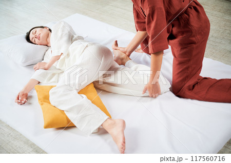 Masseuse making thai yoga massage. Cropped view of woman therapist doing traditional massage treatment, stretching female patient's body. Client lying on mat, enjoying hand massage in calm atmosphere. 117560716