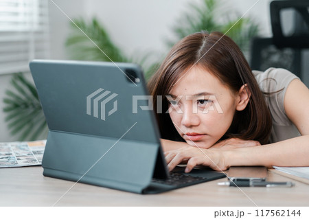 A young woman rests her head on her arms while working on a tablet in a home office, appearing tired and bored. A young woman rests her head on her arms while working on a tablet in a home office, appearing tired and bored. 117562144