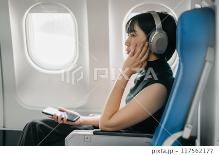 A woman wearing headphones and holding a smartphone, seated by the window on an airplane, enjoying in-flight entertainment. 117562177