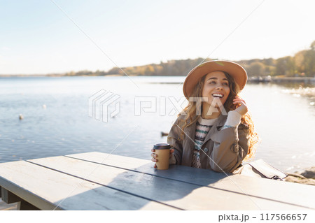Smiling woman in hat on terrace with phone enjoys sunny autumn weather. Concept of relaxation, rest. Smiling woman in hat on terrace with phone enjoys sunny autumn weather. Concept of relaxation, rest. 117566657