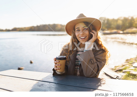 Smiling woman in hat on terrace with phone enjoys sunny autumn weather. Concept of relaxation, rest. Smiling woman in hat on terrace with phone enjoys sunny autumn weather. Concept of relaxation, rest. 117566658