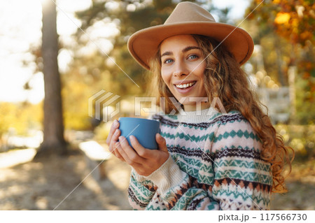 Woman holds thermos and drinks tea in sunny autumn forest. Vacation, travel, lifestyle concept. 117566730
