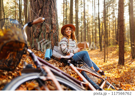 Stylish woman with a book near tree enjoys autumn weather in the park on the background of the bike. Stylish woman with a book near tree enjoys autumn weather in the park on the background of the bike. 117566762
