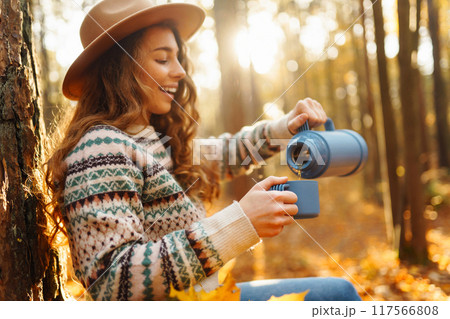 Woman traveler in hat sits near tree with thermos, enjoys autumn nature. Concept of nature. 117566808
