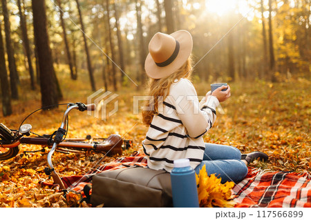 Hiker sitting on picnic blanket, drinking hot drink in autumn forest. Woman resting in nature. 117566899