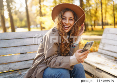 Young smiling woman sitting on bench in autumn park using phone in stylish clothes happy mood. 117566919