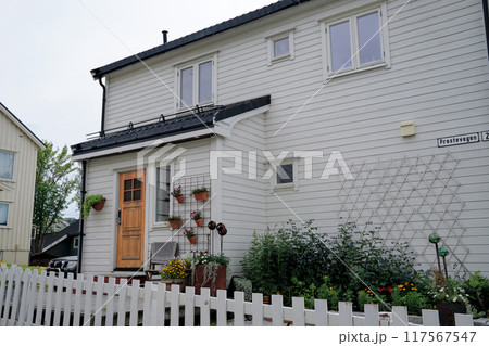 A charming white wooden house with a welcoming porch, featuring a wooden front door, potted plants, and a cozy seating area. A white picket fence and lush garden enhance the inviting atmosphere. 117567547