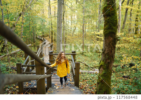 Woman in a yellow coat with backpack walks along wooden staircase, enjoying autumn scenery in forest 117568448