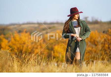 Beautiful stylish girl walks in the autumn park. The girl is dressed in a green coat and a red hat. 117569364