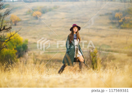 Beautiful stylish girl walks in the autumn park. The girl is dressed in a green coat and a red hat. 117569381