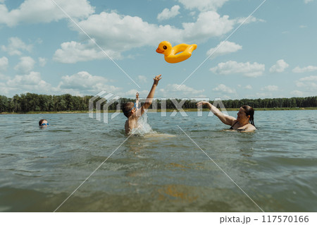 Mother and her children are enjoying a summer day at the lake, throwing an inflatable duck float. The bright sun and clear water make for a perfect vacation setting Mother and her children are enjoying a summer day at the lake, throwing an inflatable duck float. The bright sun and clear water make for a perfect vacation setting 117570166