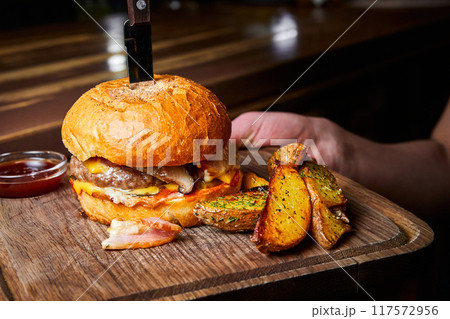 Burger with beef cutlet, a wheat bun and cheese, with a knife stuck into it, served with baked potatoes 117572956