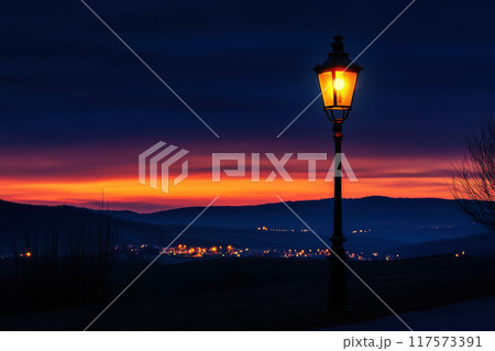 Bright street lamp shines on a street at twilight, casting a warm glow on the foggy urban landscape, with raindrops glistening in the light 117573391