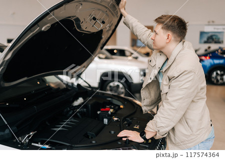 Side view of pensive male customer checking motor of new luxury car under hood in auto dealership before purchasing. Young man walking in modern auto showroom and choosing vehicle to buy or rent. Side view of pensive male customer checking motor of new luxury car under hood in auto dealership before purchasing. Young man walking in modern auto showroom and choosing vehicle to buy or rent. 117574364