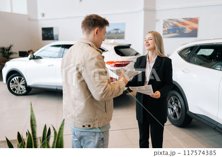 Professional blonde female car dealer in suit giving paper documents, explaining buying new car to young man at modern showroom. Concept of choosing and buying new auto in dealership office. 117574385