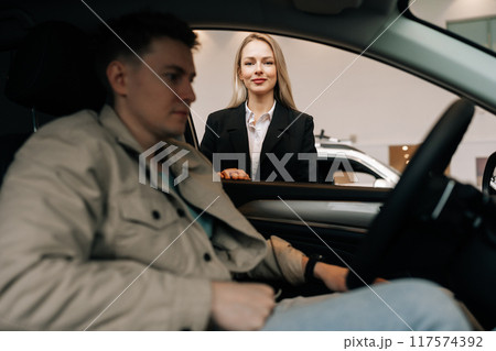 View through side window of car to professional sales female manager in suit talking to male client sitting in car in showroom choosing automobile holding steering wheel. Concept of buying new auto. 117574392