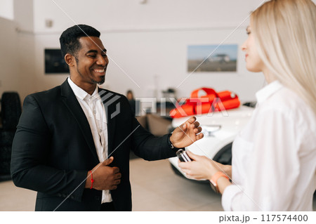 Side view of cheerful smiling female buyer getting car key and congratulation from black auto dealer male while buying new auto in dealership. Woman car owner during handing over keys at showroom. Side view of cheerful smiling female buyer getting car key and congratulation from black auto dealer male while buying new auto in dealership. Woman car owner during handing over keys at showroom. 117574400