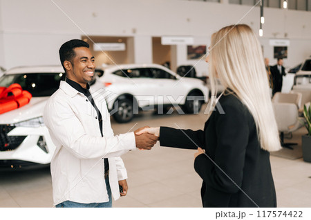 Happy cheerful female dealer and African-American customer male shaking hands in car dealership. Cheerful black young male taking key to bought car and sealing deal with handshake. 117574422
