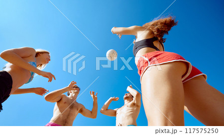 Bottom view image of four sportive young people, friends in motion, cheerfully playing beach volleyball under clear blue sky. 117575250